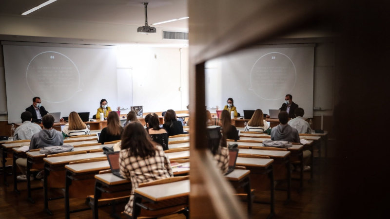 Imagem mostra sala de aula com alunos, mulheres e homens, de costas.